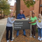 One professor and three students stand around a building sign that reads "Meyerhoff Chemistry Building" outside on a fall day, backed by a walkway and brick buildings