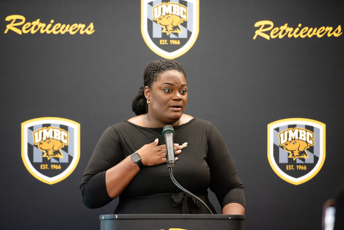 A woman, Tiffany Tucker, stands at a podium in a black dress with a gold UMBC pin, hand to her heart, in front of a UMBC Retrievers step and repeat 