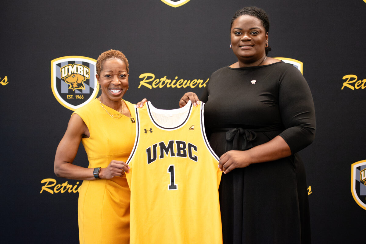 UMBC's president Valerie Sheares Ashby and UMBC's athletic director Tiffany turner stand in front of a UMBC Retrievers step and repeat with a UMBC basketball jersey #1 between them 