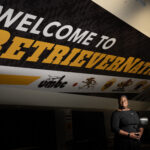 UMBC's AD Tiffany Turner stands in a black dress in front of massive poster spanning slanted ceiling that reads "welcome to retriever nation"