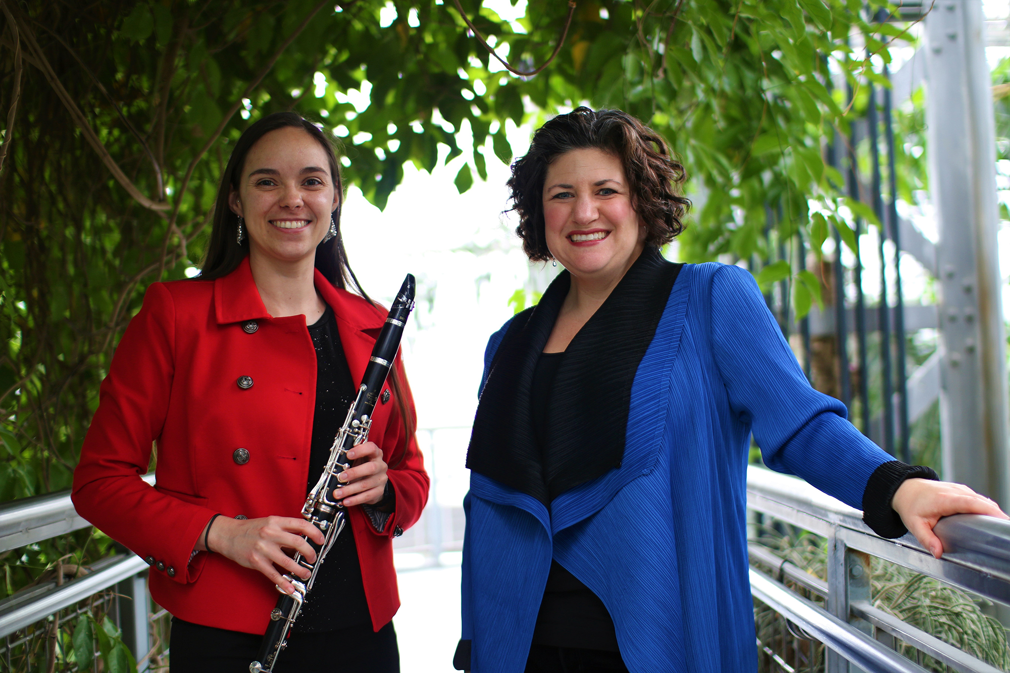 Two women smile at the camera. On the left, a woman holds a flute and wears red. On the right, the woman is dressed in blue and black.