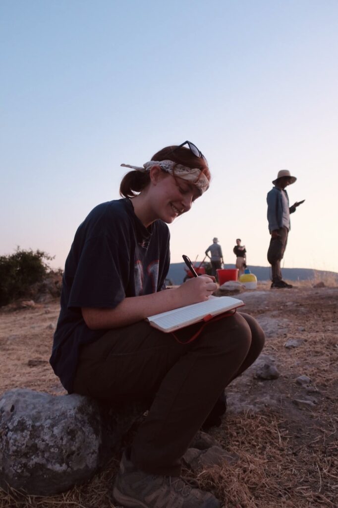 An archaeological student kneels down to document in a notebook with other students working in the background.