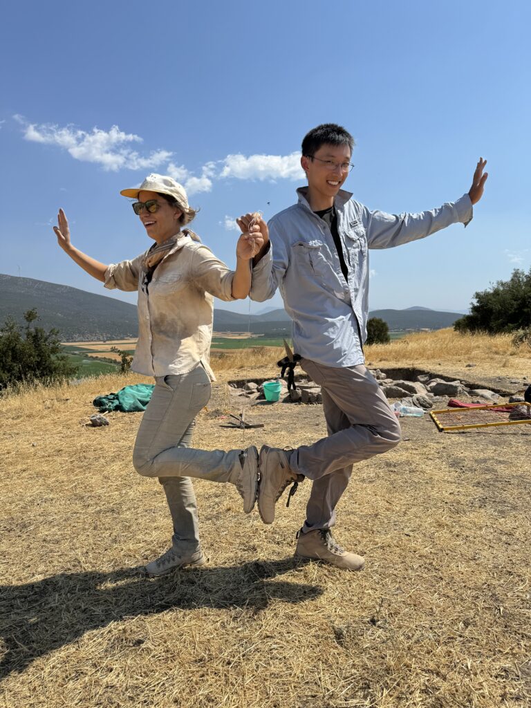 Robert Barry and crew mate wear khaki clothes stand with their back to each other with their legs raised and bend places the bottoms of their shoes against each other  while they hold hands and raise one up to the side