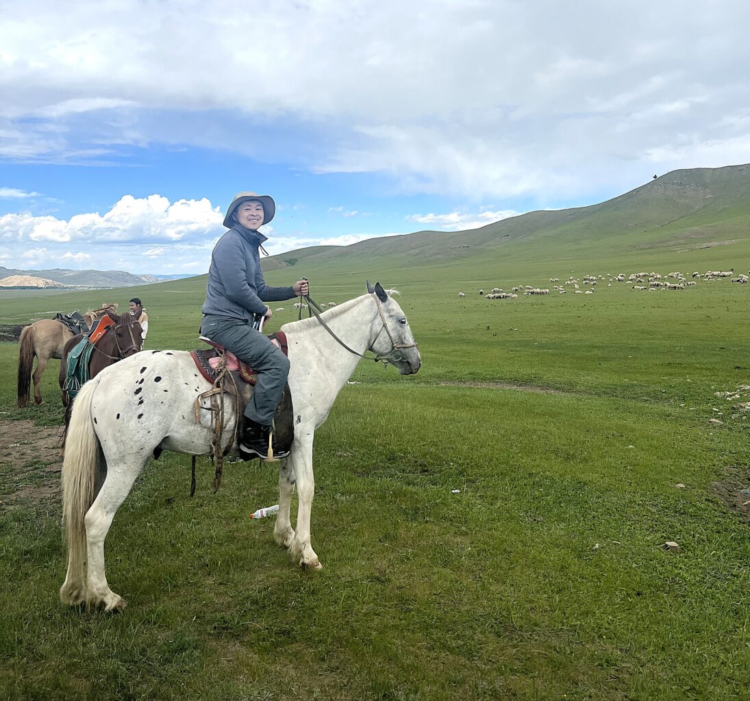 A man in Mongolia wears grey pants and a grey sweater as he sits on top of a white horse with an expansive field and hills in the background along with another person who stands next to two other horses.