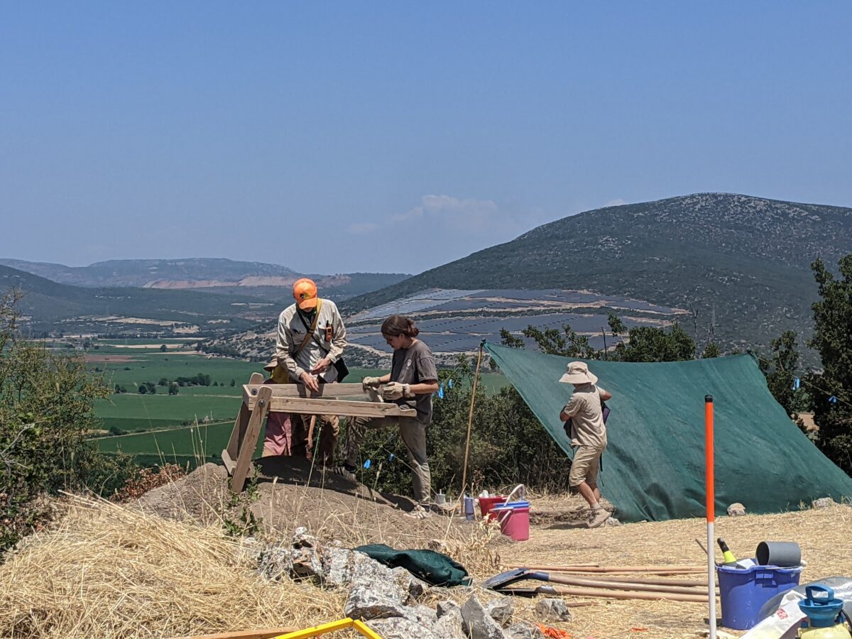 Two archeology students use a stand up mesh screen to sift soil for artifacts in Greede