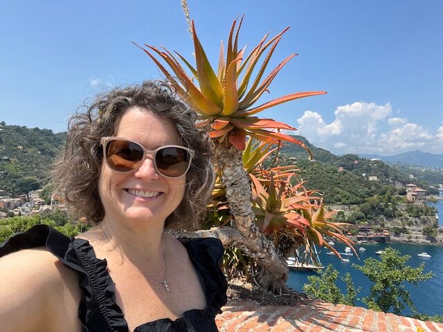 Susan McDonough, a person with neck-length brown and white wavy hear wearing a black ruffled tank top stands in front of an orange and yellow palm tree with a view of an Italian seaport in the background.