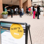 outdoor pedestrian corridor with a large group of professionally dressed students walking across; in foreground an easel with poster advertising Summer Undergraduate Research Fest that reads "SURF's UP 2024" at the top