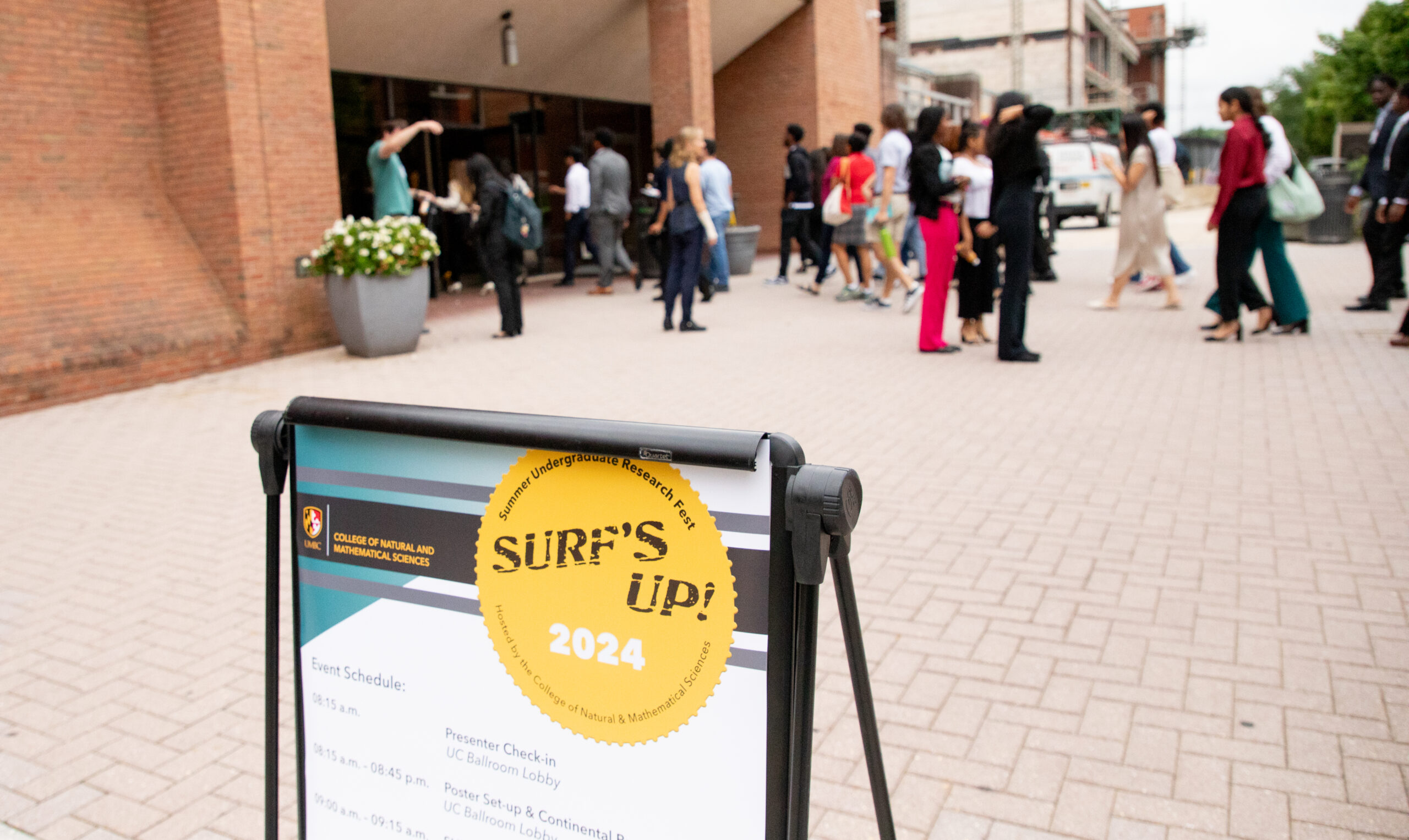 outdoor pedestrian corridor with a large group of professionally dressed students walking across; in foreground an easel with poster advertising Summer Undergraduate Research Fest that reads "SURF's UP 2024" at the top