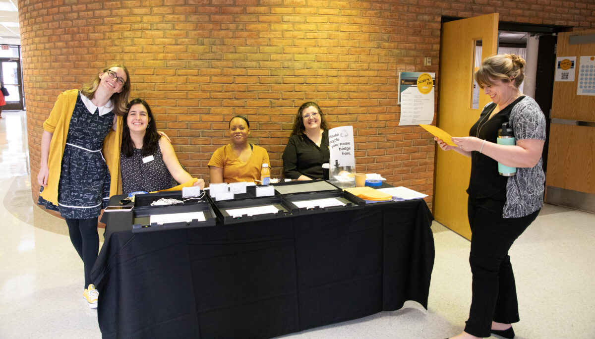 Four people sit at a table with a black tablecloth over it, one stands next to it, in front of a brick wall. All are dressed in black and gold and smiling.