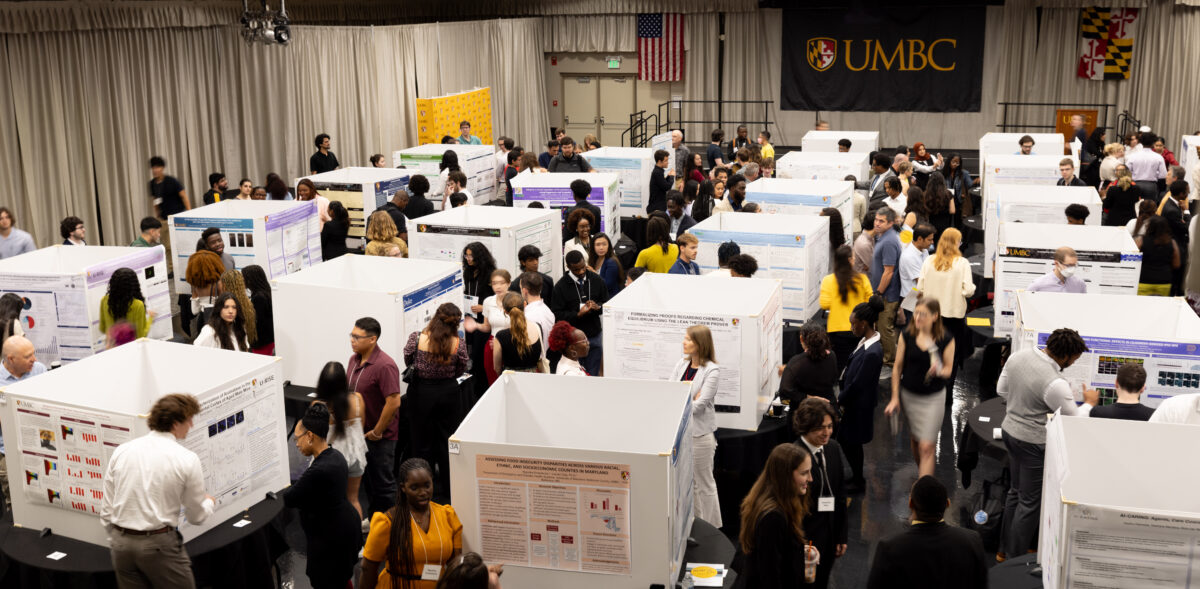 An overhead view of a packed exhibition hall including square-shaped displays of research posters on tables (one poster on each side of the square) and many people walking around and having conversation 