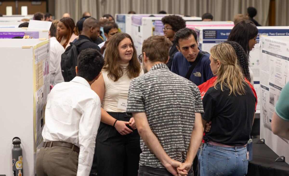 Members of the UMBC community and visitors interact with student researchers in a packed exhibition hall full of animated people and research posterboards on tables.