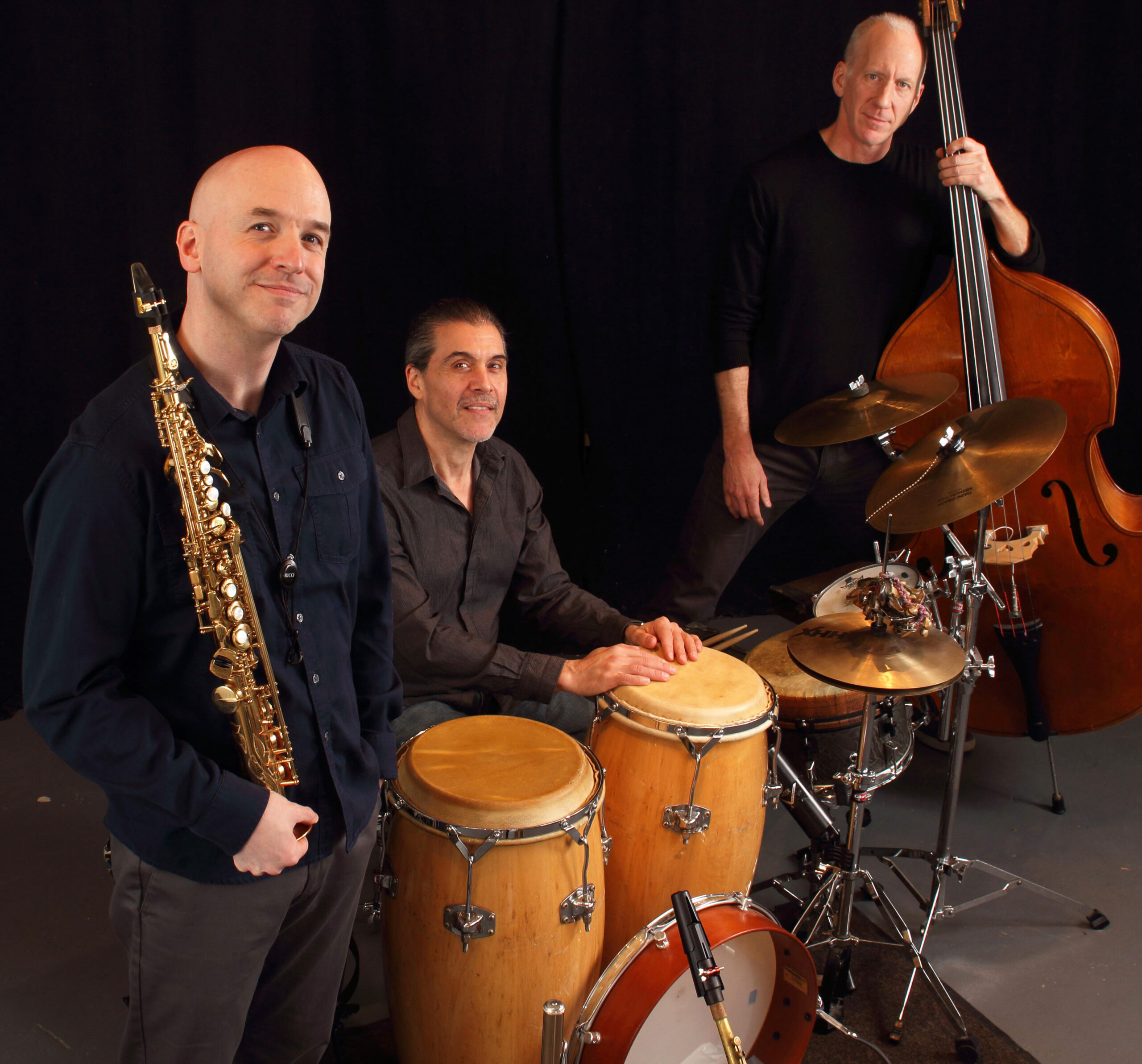 Against a black background, three people pose with musical instruments — left to right, saxophone, drums and double bass.