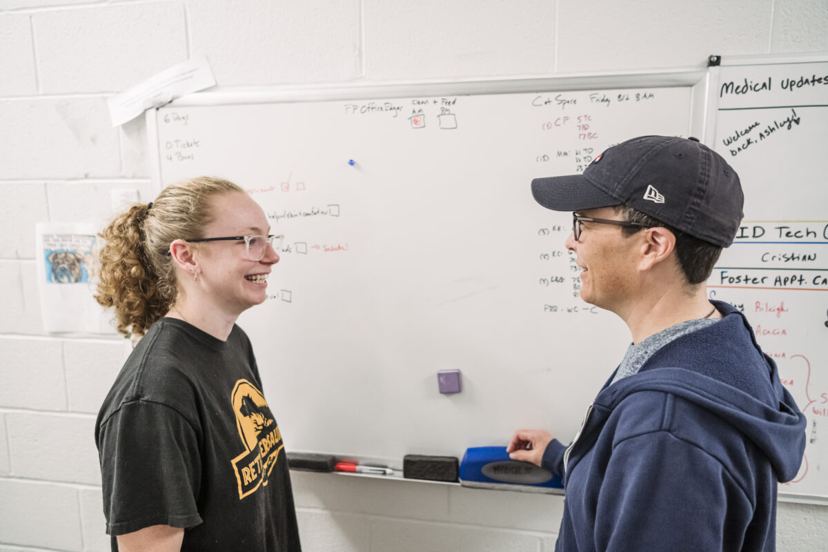 A student with curly hair and glasses stands at a white board with a staff member (one helping her with her vet training) wearing a baseball cap and glasses.