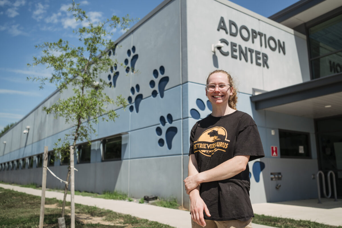 A girl with curly hair in a ponytail and a black t-shirt stands in front of a large building with paw prints on the Side and "Adoption Center"