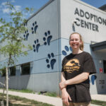 A girl with curly hair in a ponytail and a black t-shirt stands in front of a large building with paw prints on the Side and "Adoption Center"