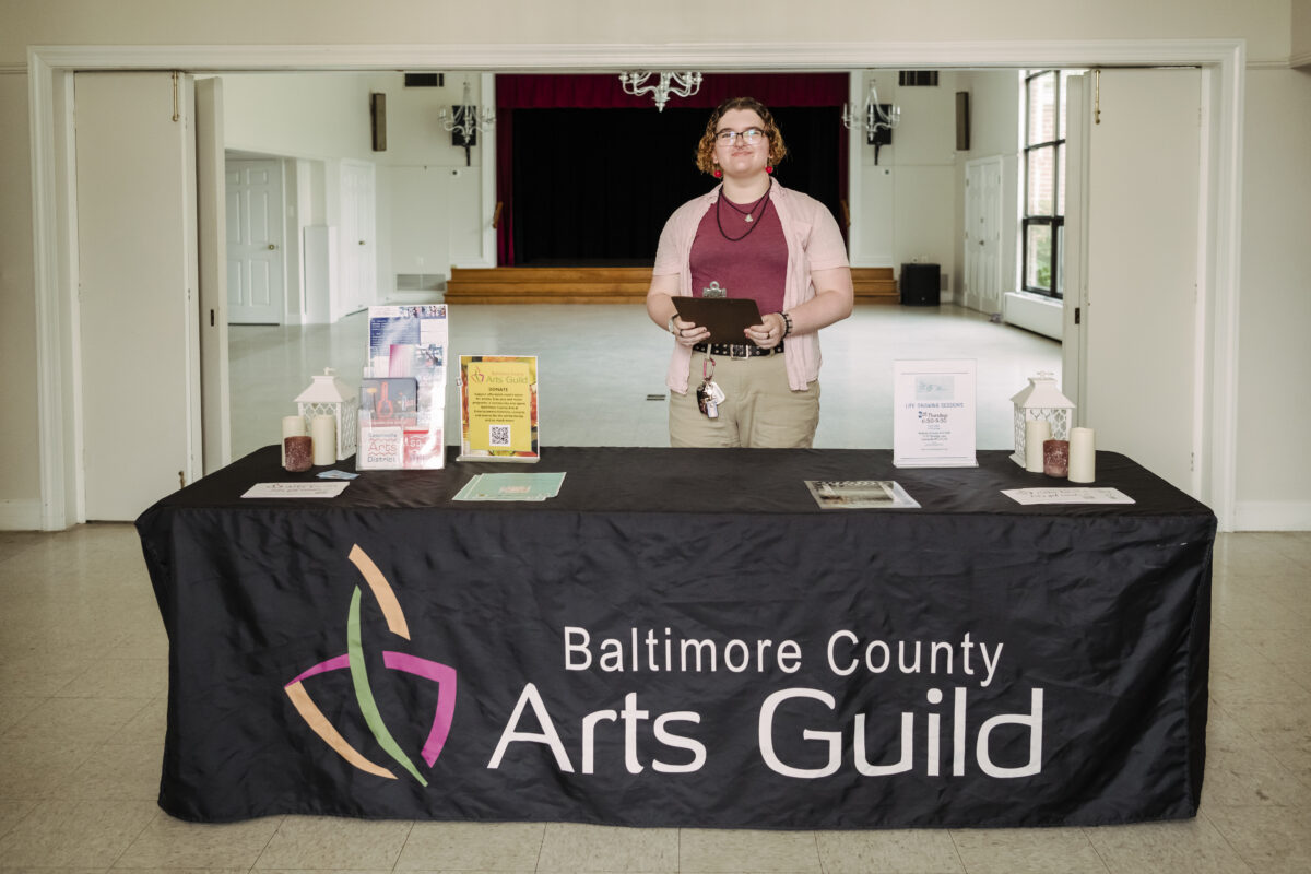 a student stands behind a table that says Baltimore County Arts Guild as part of her internships duties