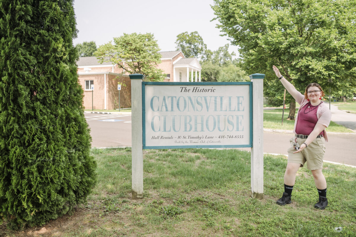 In an outdoor setting, a person, smiling, gestures toward a sign on the left that says Catonsville Clubhouse.