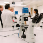 a white microscope on a white lab bench in a bright laboratory; two people in lab coats and two without have a conversation in the background