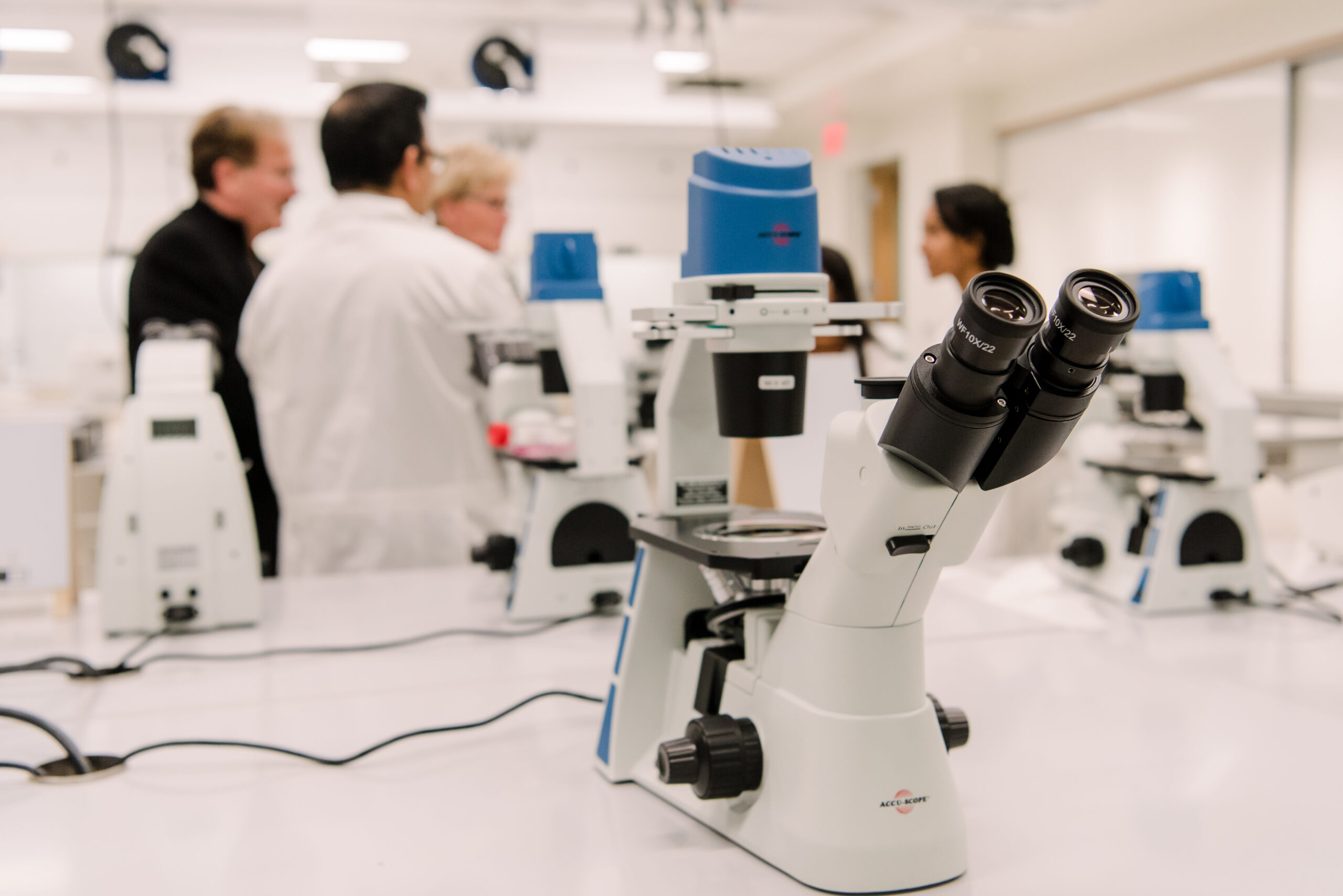 a white microscope on a white lab bench in a bright laboratory; two people in lab coats and two without have a conversation in the background