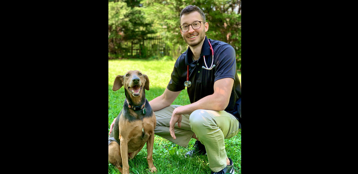 a vet crouches down with a dog, sitting on the grass on a sunny day