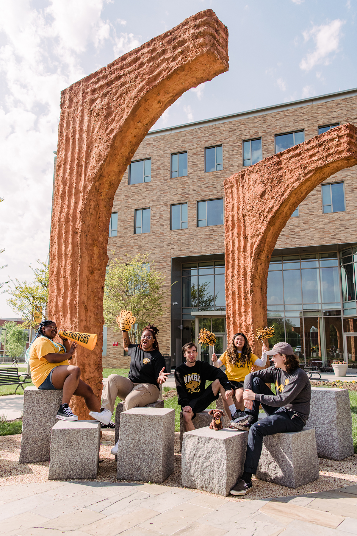 A diverse group of UMBC students, wearing black and gold spirit shirts, sitting under sculptural archways on campus