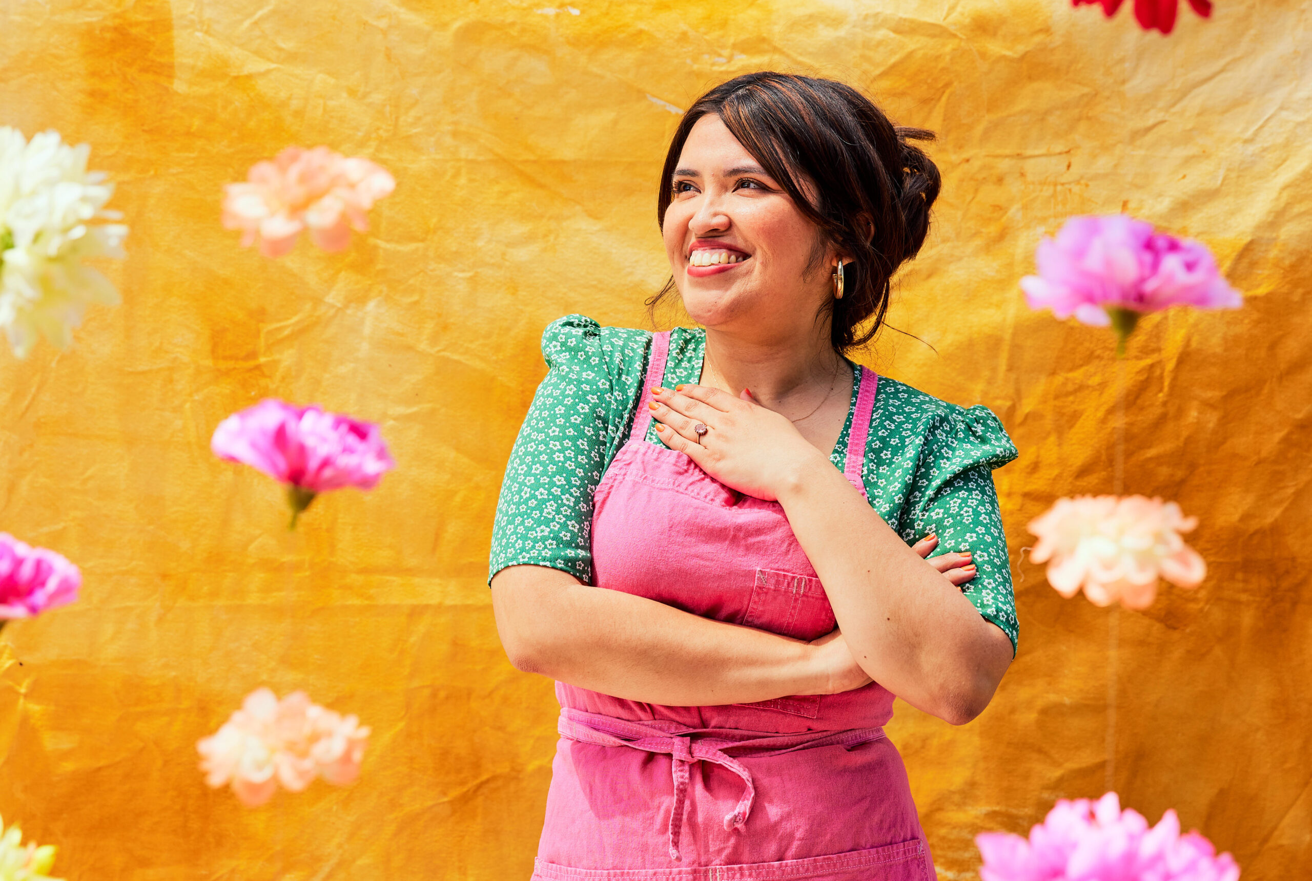 Against a colorful background, a woman with dark hair, wearing a green top and magenta overalls, smiles and looks into the distance.