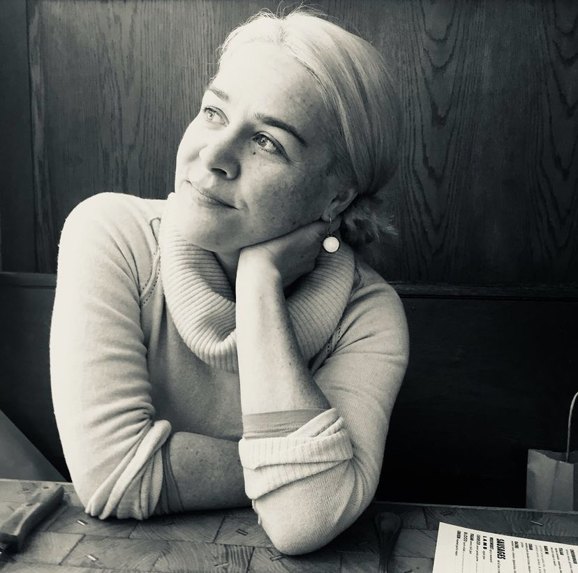 In a black and white image, a woman seated at a table wears a sweater and looks into the distance.