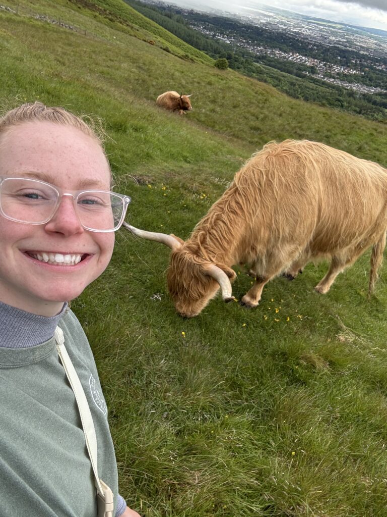 A girl in a gray sweatshirt and glasses smiles for a selfie with a Highland cow