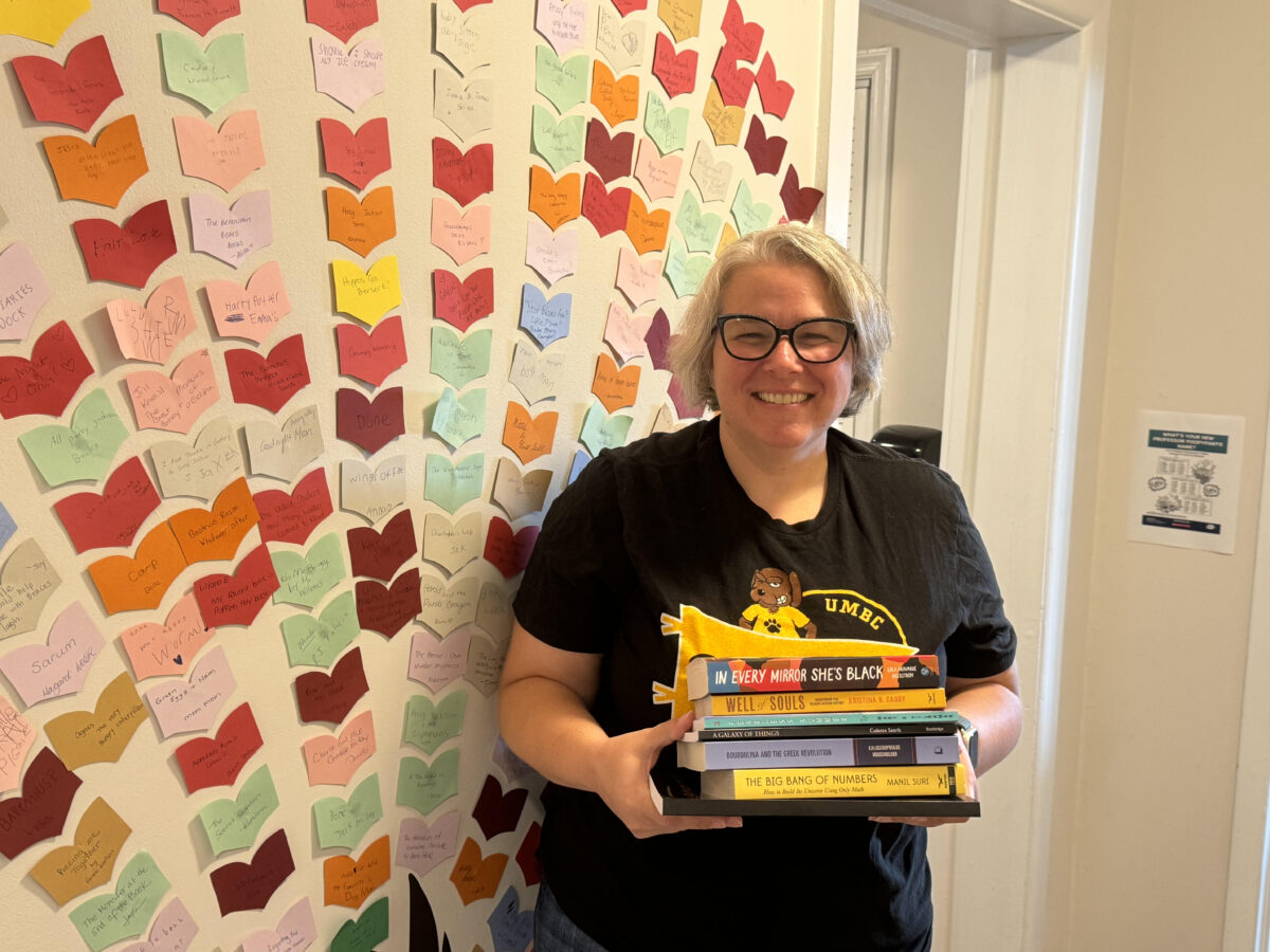 Jennifer Matthews '02, owner of the independent bookstore Growing Minds in Catonsville, stands against a wall holding a stack of books.