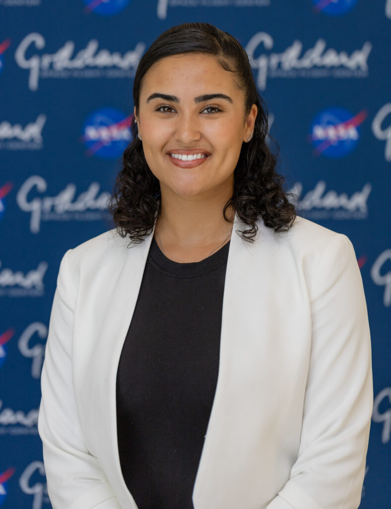 Woman in white coat and black shirt stands in front of blue background with NASA Goddard logos, smiles at camera.