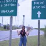 Woman stands between two signs near a road. One sign reads "International Space Station, On-ramp" another heads "Moon Ahead."