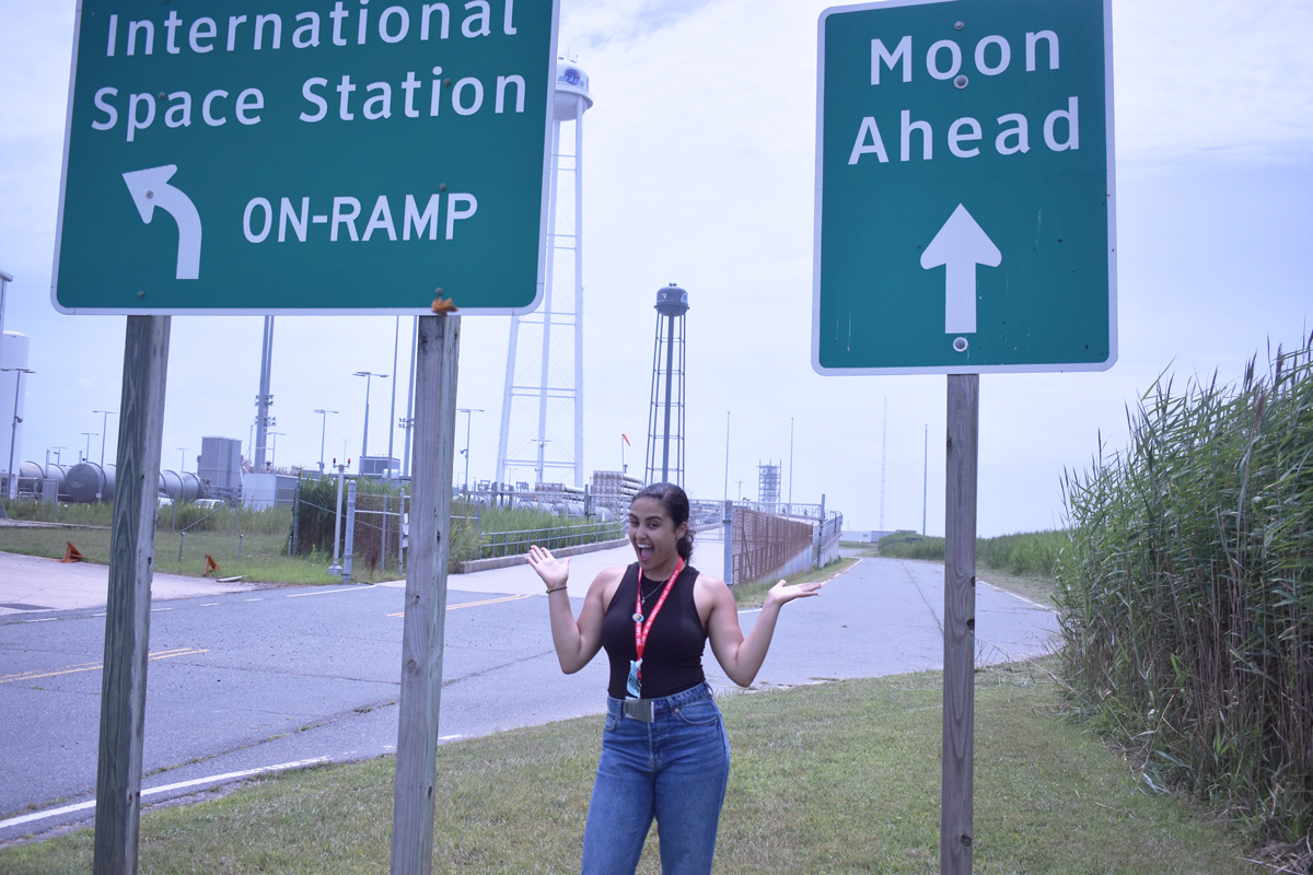 Woman stands between two signs near a road. One sign reads "International Space Station, On-ramp" another heads "Moon Ahead."