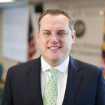 A headshot of Josh Michael, an adult man, wearing a black dress jacket, a green and block polka dotted tie, and white dress collar shirt