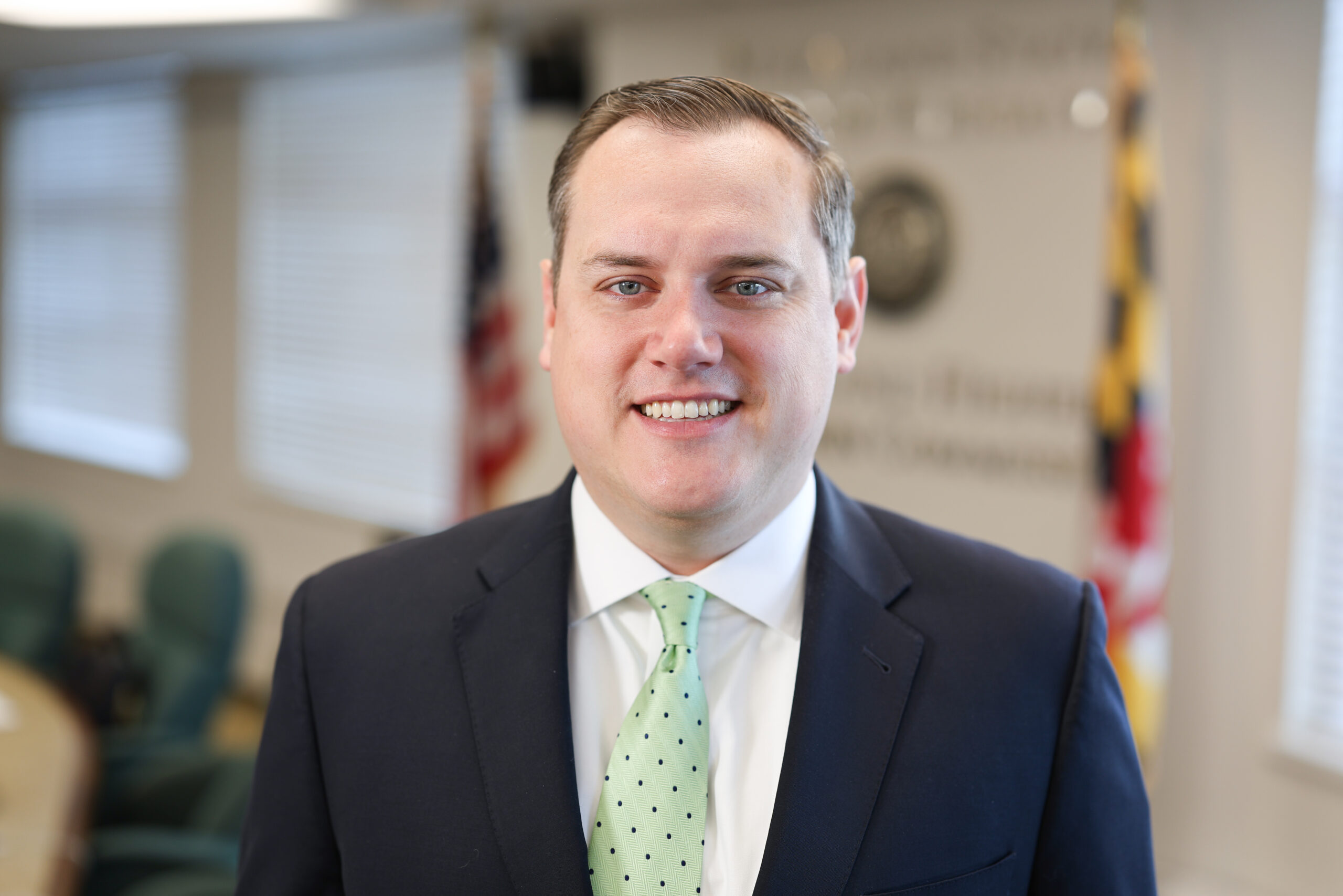 A headshot of Josh Michael, an adult man, wearing a black dress jacket, a green and block polka dotted tie, and white dress collar shirt