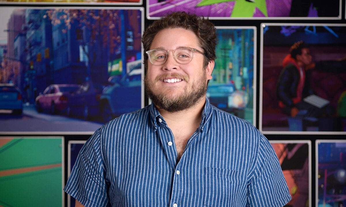 Against a background of artistic images, a person with short brown hair and a beard wears glasses and a blue shirt, and smiles at the camera.