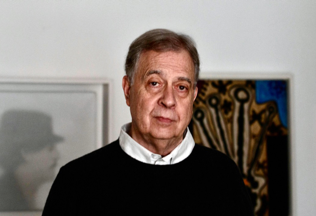 A white man with short dark hair, wearing a white shirt and dark sweater, stands against to paintings and looks at the camera.