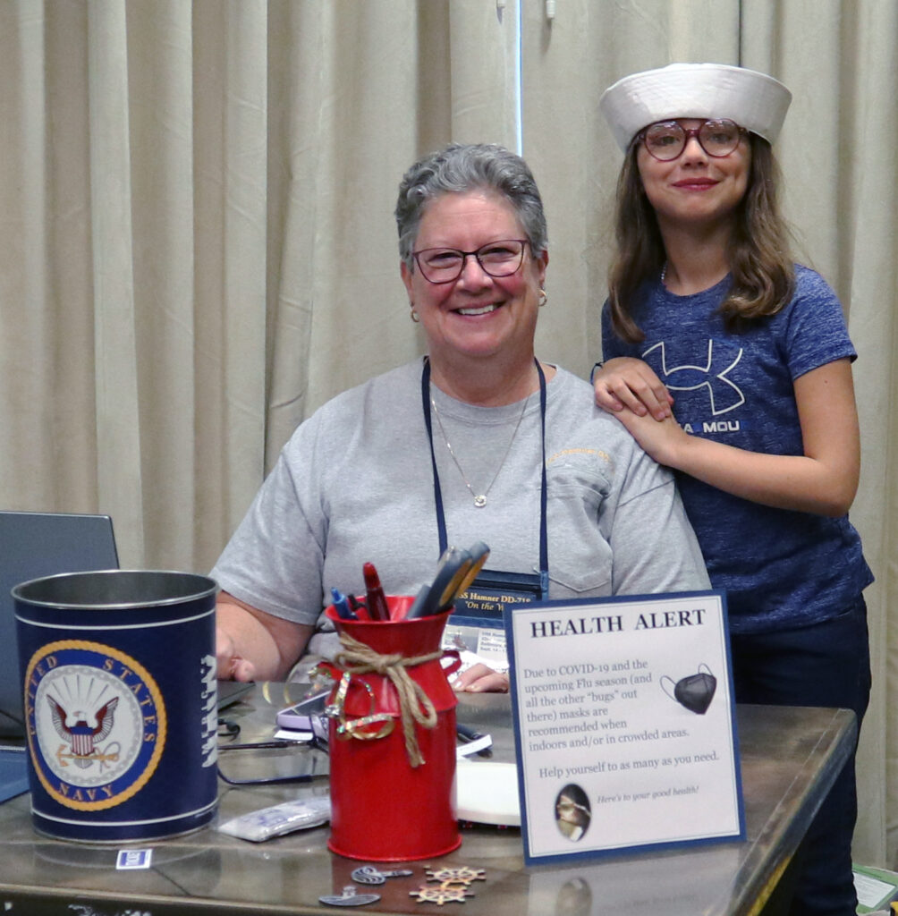 an older woman and a younger woman in a navy cap pose behind a desk welcome naval veterans to a reunion