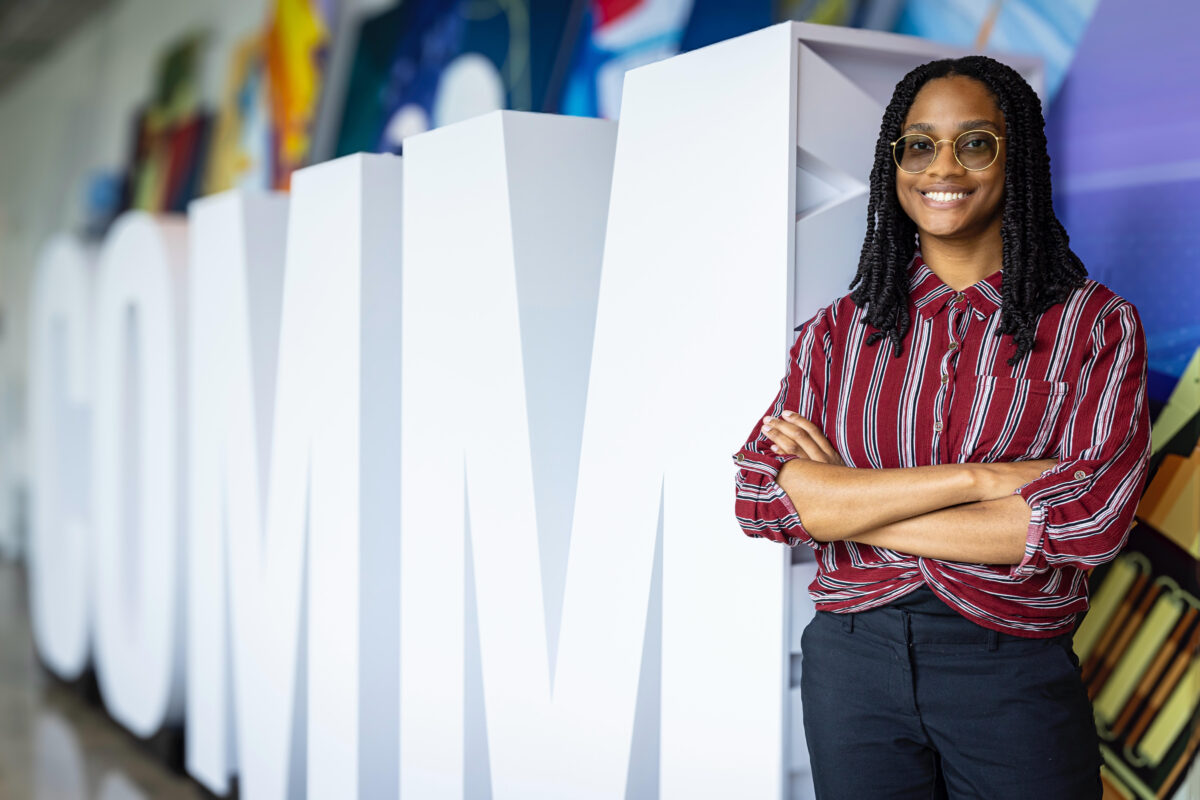 a young woman stands with glasses and her arms crossed in front of large cut out letters that spell COMM