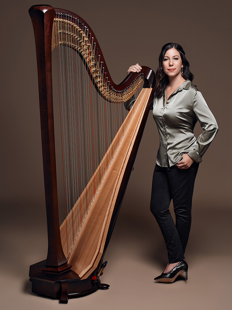 Against a brown background, a person wearing a silver top poses with a harp.