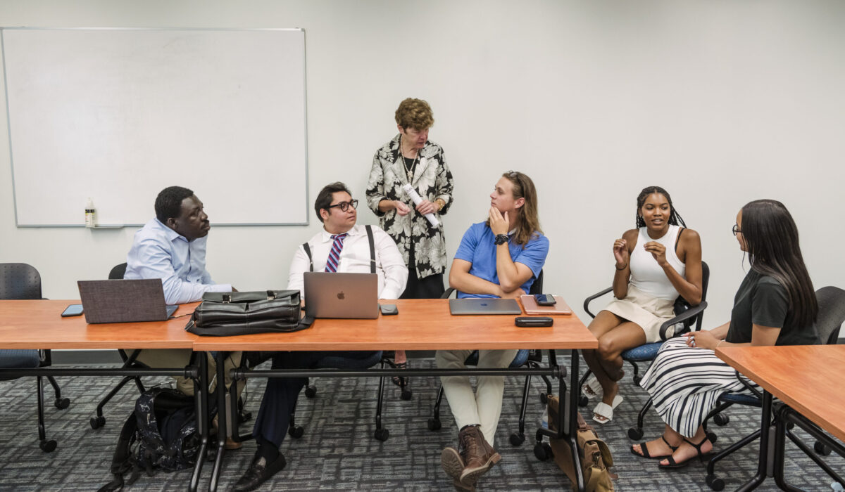 A professor stands to speak with a student who is seated at a table