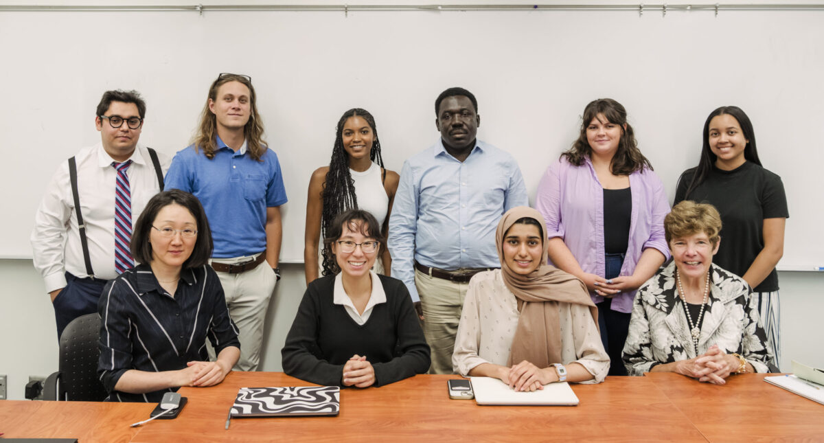 A group of eight college students stand behind two faculty and two students who are seated at a table