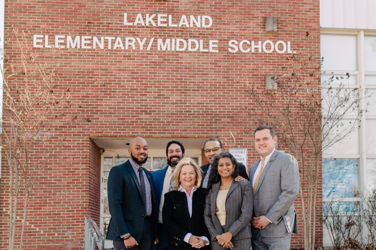 A group of six educators stand in a group in business attire in front of a red brick school building with the school name posted in white letters