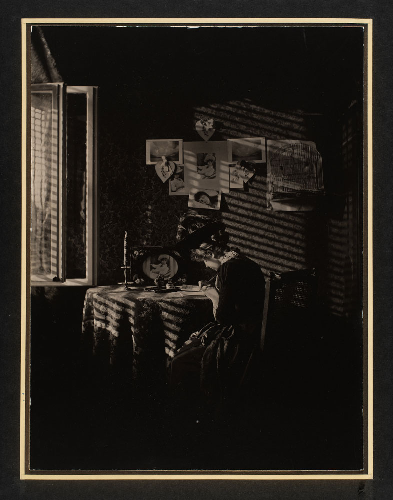 In a dark, sepia-tone photograph by Alfred Stieglitz, a woman writes a letter at a table.