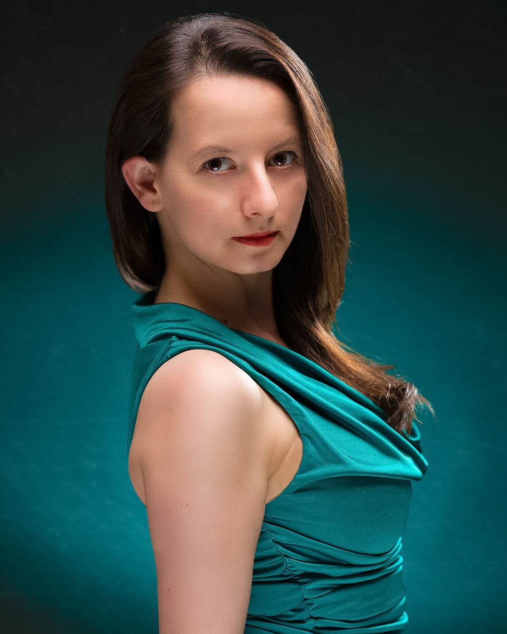 Against a turquoise background, a person with a medium light skin tone and dark hair, wearing a turquoise top, turns her head to face the camera.