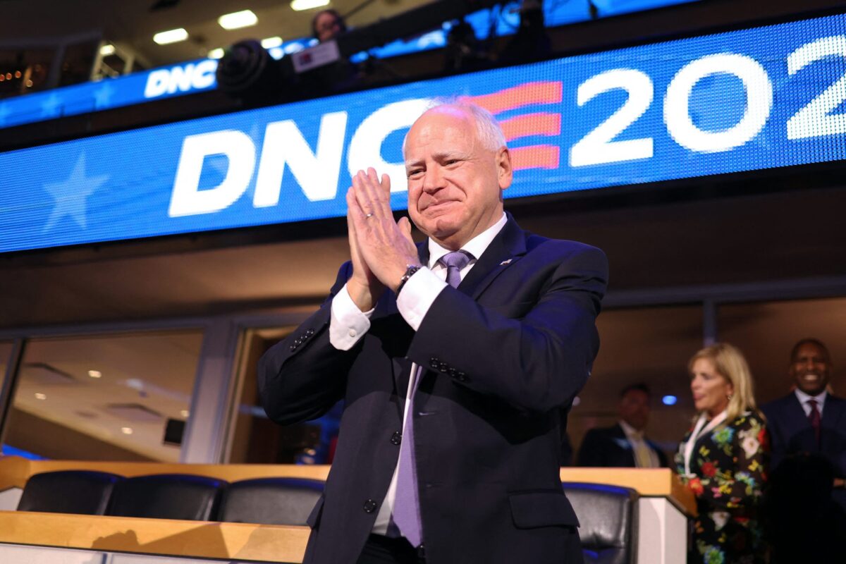 A U.S. vice presidential candidate stands with his hands raised in prayer with the white and blue DNC 2024 digital sign behind them