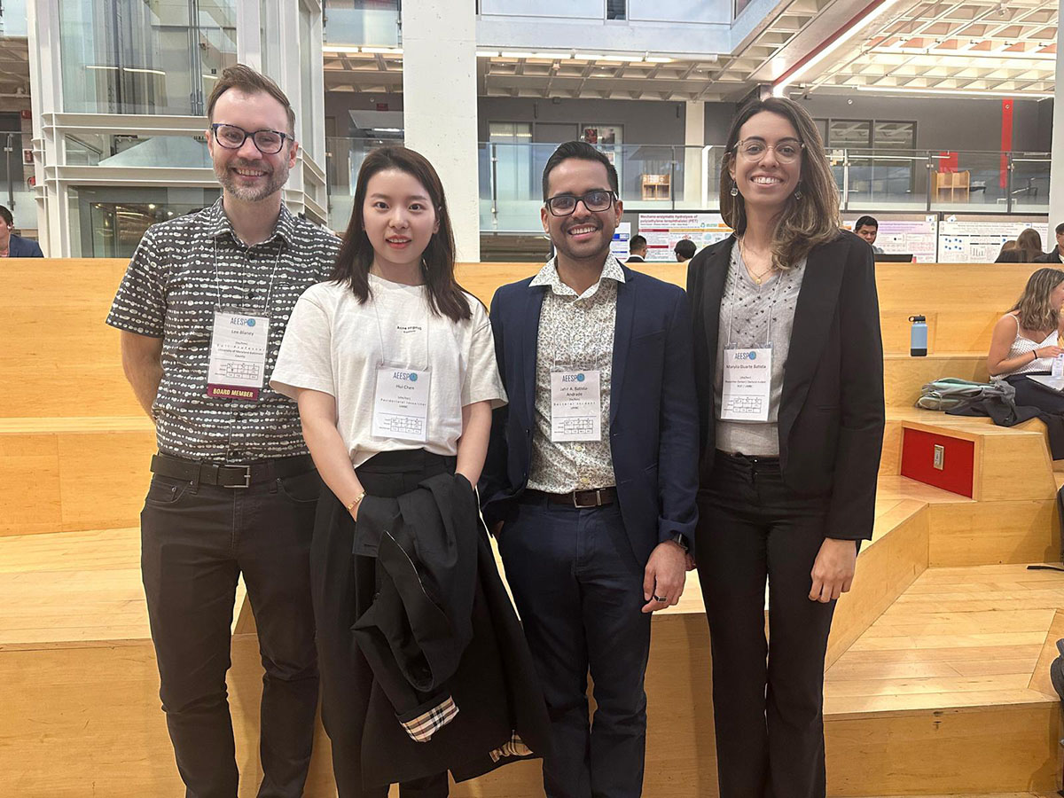 Four people wearing conference badges stand in large room and smile at camera.