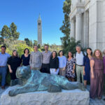 A large group of people gather outside behind a metal statue of a bear. A tall tower is in the distance.