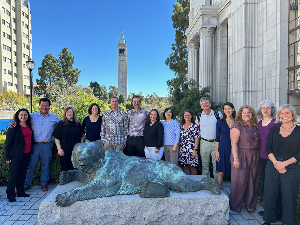 A large group of people gather outside behind a metal statue of a bear. A tall tower is in the distance.