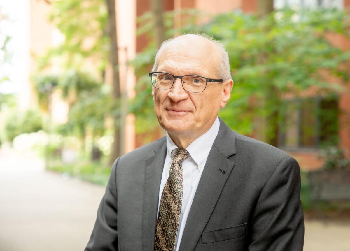 portrait of smiling man wearing suit on walkway, brick buildings and green trees in background