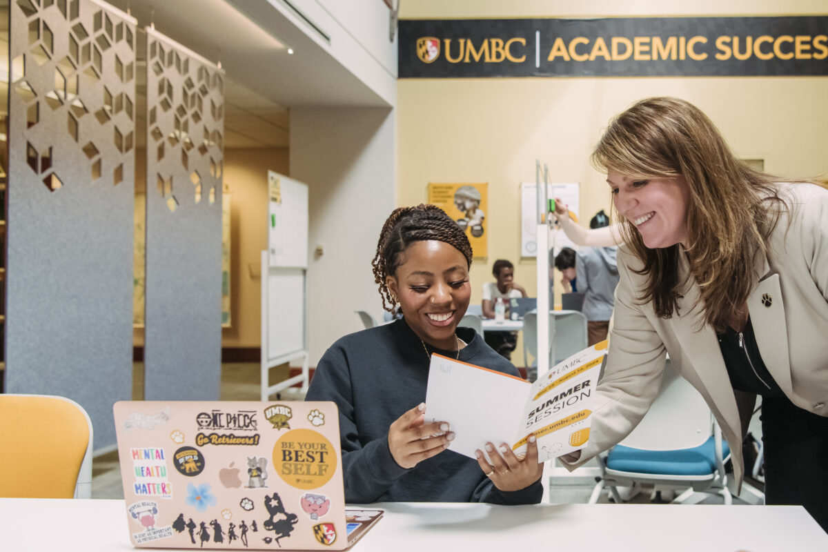 student seated at a table with a laptop in front of her looking at a booklet that reads "summer session" with a staff member who is standing nearby and leaning over pointing to the booklet